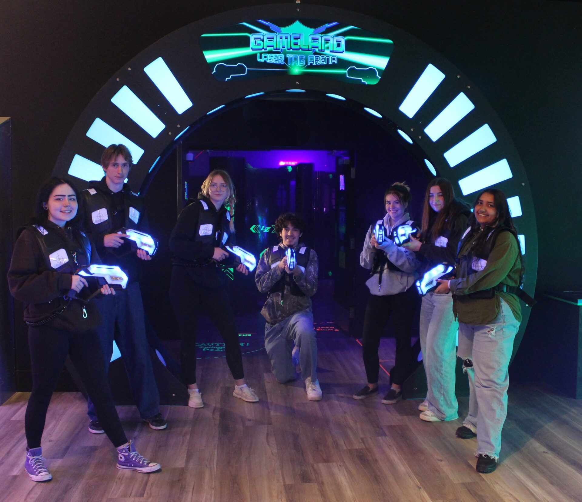 A large circular arch with neon lights and the Gameland Laser Tag and Arcade logo against a black wall. Behind the arch, a door opens into a laser tag arena - black walls in a scattered confirguration are painted with orange and yellow arrows. Under the arch, a group of 7 teens wear laser tag vests - black with glowing purple and blue lights - and hold laser tag guns - about the length of their forearms and glowing blue. They are in an at ease position and smiling at the camera.